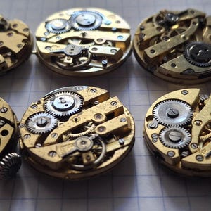 Op de afbeelding: Een verzameling vintage uurwerkonderdelen, met ingewikkelde tandwielen, veren en andere mechanische componenten. De messingkleurige onderdelen zijn gerangschikt op een rasteroppervlak, wat de gedetailleerde vakmanschap benadrukt. Ideaal voor restauratie of knutselprojecten.