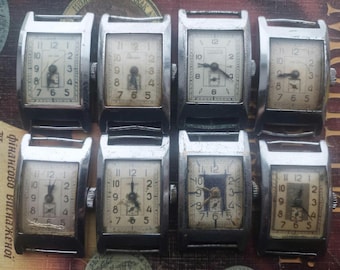 Può includere: Una collezione di otto orologi da polso rettangolari vintage con casse argentate e quadranti bianchi sporchi. I quadranti mostrano numeri e lancette nere. Gli orologi sono disposti su una superficie strutturata.