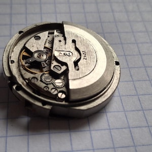 K&ouml;nnte beinhalten: Nahaufnahme eines zerlegten Uhrwerks, das komplizierte Zahnr&auml;der, Federn und andere mechanische Komponenten zeigt. Die Metallteile sind silber- und graufarben, mit der Nummer "2427" auf dem Zifferblatt. Das Uhrwerk liegt vor einem karierten Hintergrund.