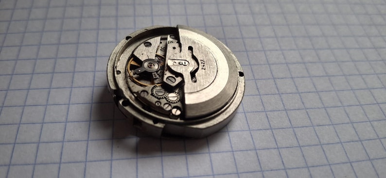 K&ouml;nnte beinhalten: Nahaufnahme eines zerlegten Uhrwerks, das komplizierte Zahnr&auml;der, Federn und andere mechanische Komponenten zeigt. Die Metallteile sind silber- und graufarben, mit der Nummer "2427" auf dem Zifferblatt. Das Uhrwerk liegt vor einem karierten Hintergrund.