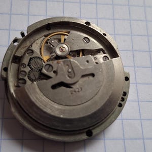 May include: A detailed view of a watch movement, revealing its internal components. The silver-toned metal parts, including gears and springs, are arranged within the watch casing. The intricate mechanism is displayed, highlighting the precision engineering.