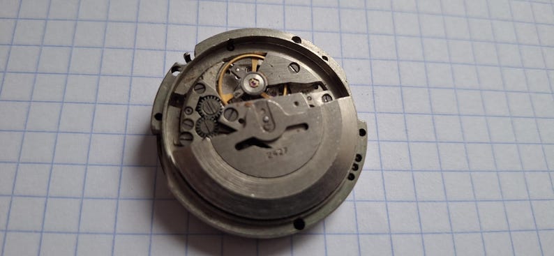 K&ouml;nnte beinhalten: Nahaufnahme eines zerlegten Uhrwerks. Die silberfarbenen Metallkomponenten, darunter Zahnr&auml;der und Federn, sind zu sehen. Das Innenleben der Uhr wird gezeigt und zeigt das komplizierte Design und die Handwerkskunst des Zeitmessers.