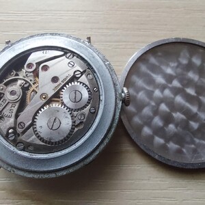 Puede incluir: Primer plano de un reloj vintage con la tapa trasera abierta. El mecanismo del reloj es visible, con engranajes, tornillos y la inscripci&oacute;n "SWISS MADE". El reloj tiene una caja plateada y una tapa trasera texturizada.