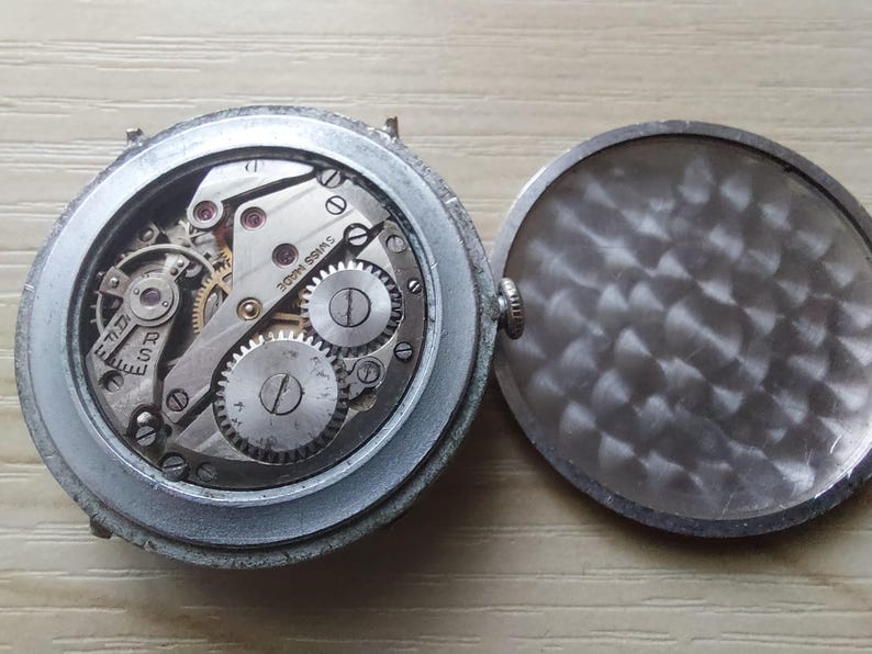 Puede incluir: Primer plano de un reloj vintage con la tapa trasera abierta. El mecanismo del reloj es visible, con engranajes, tornillos y la inscripci&oacute;n "SWISS MADE". El reloj tiene una caja plateada y una tapa trasera texturizada.