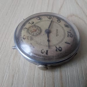Puede incluir: Reloj de pulsera Zenith vintage con caja plateada y esfera crema. El reloj presenta n&uacute;meros ar&aacute;bigos negros, una subesfera y agujas negras. Tiene una peque&ntilde;a corona y est&aacute; sobre una superficie marr&oacute;n claro.