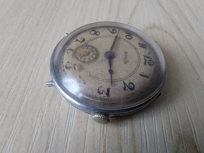 Puede incluir: Reloj de pulsera Zenith vintage con caja plateada y esfera crema. El reloj presenta n&uacute;meros ar&aacute;bigos negros, una subesfera y agujas negras. Tiene una peque&ntilde;a corona y est&aacute; sobre una superficie marr&oacute;n claro.
