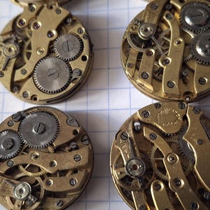 Op de afbeelding: Vier vintage uurwerken met zichtbare tandwielen en ingewikkelde mechanische componenten. De horlogeonderdelen zijn goudkleurig met zilverkleurige tandwielen, gerangschikt op een witte rasterachtergrond. Close-up.
