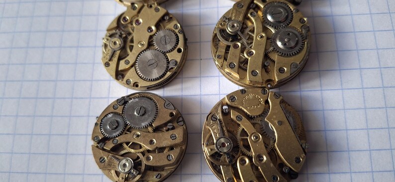 Op de afbeelding: Vier vintage uurwerken met zichtbare tandwielen en ingewikkelde mechanische componenten. De horlogeonderdelen zijn goudkleurig met zilverkleurige tandwielen, gerangschikt op een witte rasterachtergrond. Close-up.