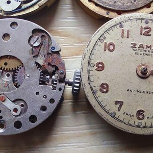 May include: Close-up of two vintage watch movements. One is a silver-toned mechanism with gears and small components. The other is a watch face with the text "ZAMA WATERPROOF 15 JEWELS" and the word "ANTIMAGNETIC".