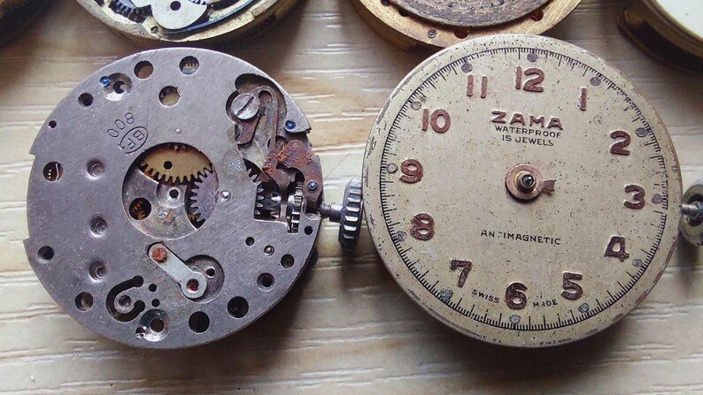 May include: Close-up of two vintage watch movements. One is a silver-toned mechanism with gears and small components. The other is a watch face with the text "ZAMA WATERPROOF 15 JEWELS" and the word "ANTIMAGNETIC".