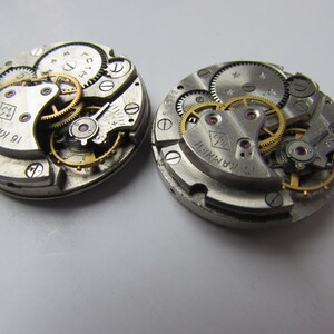 May include: Two silver watch movements with intricate gears and a gold-colored winding stem. The watch movements are labeled "16 KAMNEN" and "17 KAMNEN".
