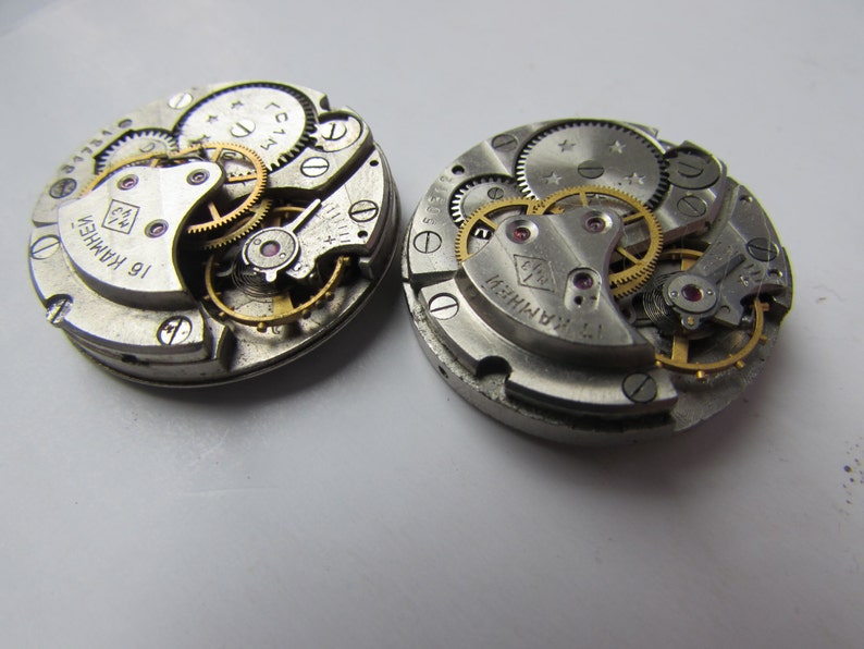 May include: Two silver watch movements with intricate gears and a gold-colored winding stem. The watch movements are labeled "16 KAMNEN" and "17 KAMNEN".
