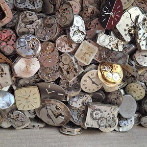 K&ouml;nnte beinhalten: Eine Sammlung von Vintage-Uhrenteilen, darunter Zifferbl&auml;tter, Zahnr&auml;der und Innenmechanismen. Die Komponenten sind in verschiedenen Formen und Gr&ouml;&szlig;en gehalten, mit einer Mischung aus Silber-, Gold- und Messingt&ouml;nen. Die Teile befinden sich in einem Karton.
