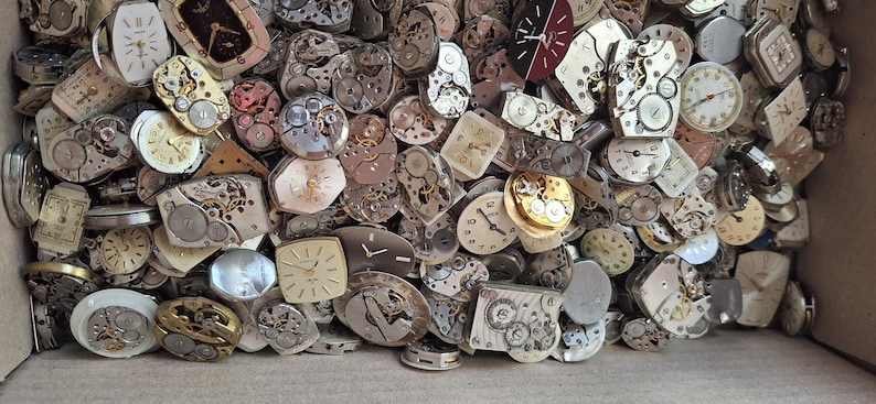 K&ouml;nnte beinhalten: Eine Sammlung von Vintage-Uhrenteilen, darunter Zifferbl&auml;tter, Zahnr&auml;der und Innenmechanismen. Die Komponenten sind in verschiedenen Formen und Gr&ouml;&szlig;en gehalten, mit einer Mischung aus Silber-, Gold- und Messingt&ouml;nen. Die Teile befinden sich in einem Karton.