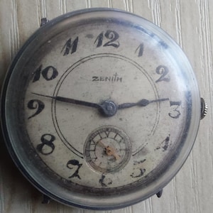 Puede incluir: Reloj de pulsera Zenith vintage con caja redonda plateada. La esfera blanquecina presenta n&uacute;meros ar&aacute;bigos negros, un subdial y agujas negras. El reloj muestra signos de edad, con un aspecto ligeramente desgastado.