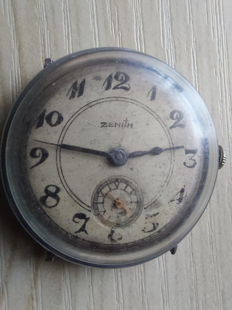 Puede incluir: Reloj de pulsera Zenith vintage con caja redonda plateada. La esfera blanquecina presenta n&uacute;meros ar&aacute;bigos negros, un subdial y agujas negras. El reloj muestra signos de edad, con un aspecto ligeramente desgastado.