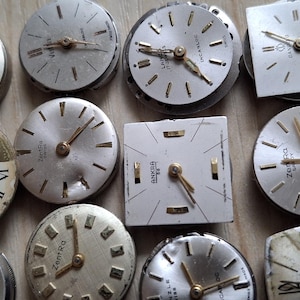 May include: A collection of vintage watch faces in various shapes, including round and square. The faces are silver and white, with gold-toned hands and markers. Some faces show Roman numerals. The watch faces are arranged on a light brown surface.