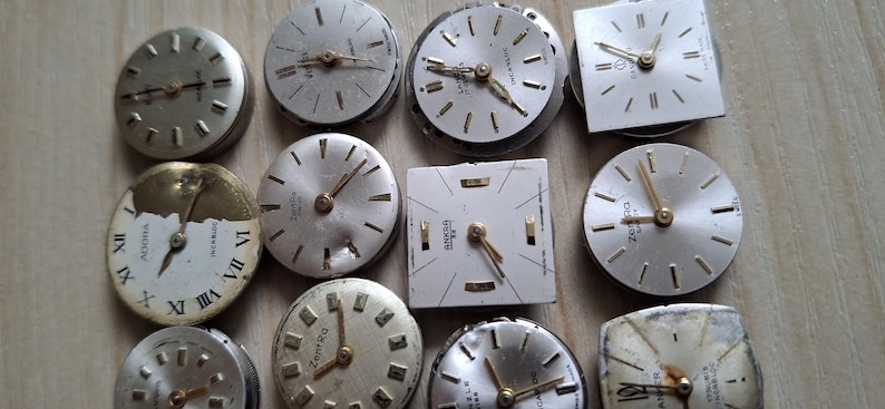 May include: A collection of vintage watch faces in various shapes, including round and square. The faces are silver and white, with gold-toned hands and markers. Some faces show Roman numerals. The watch faces are arranged on a light brown surface.
