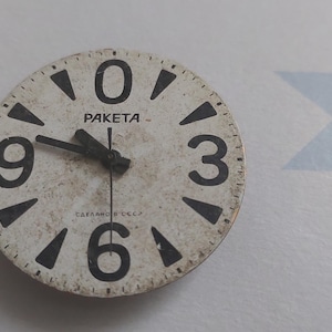 Puede incluir: Una esfera de reloj redonda y vintage con números y manecillas negras sobre un fondo crema. La palabra "PAKETA" está impresa en el centro. La hora que se muestra es aproximadamente las 9:50.