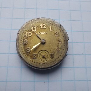 May include: A vintage watch face with a gold-toned dial and hands. The watch face has black numerals and the brand name "DOXA" is printed at the top. A small subdial is at the bottom. The watch face is set against a blue grid background.