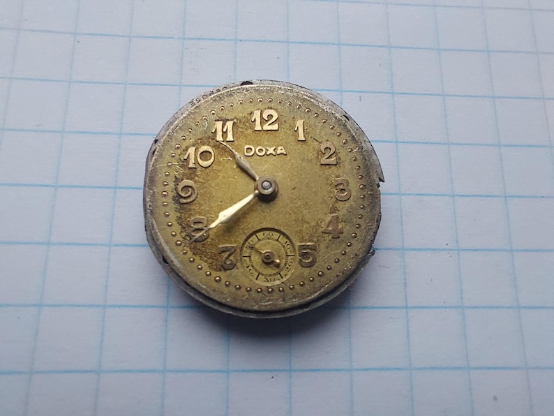 May include: A vintage watch face with a gold-toned dial and hands. The watch face has black numerals and the brand name "DOXA" is printed at the top. A small subdial is at the bottom. The watch face is set against a blue grid background.