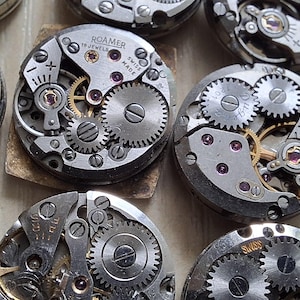 May include: Close-up of several vintage watch movements, showcasing intricate gears, springs, and jewels. The silver-toned metal components are arranged in a circular pattern, with some featuring gold accents. The image highlights the detailed craftsmanship of antique watchmaking.