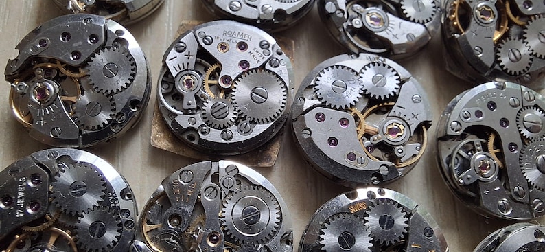 May include: Close-up of several vintage watch movements, showcasing intricate gears, springs, and jewels. The silver-toned metal components are arranged in a circular pattern, with some featuring gold accents. The image highlights the detailed craftsmanship of antique watchmaking.