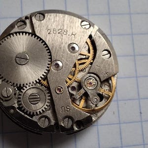 Könnte beinhalten: Nahaufnahme eines Uhrwerks mit sichtbaren Zahnrädern und komplexen mechanischen Komponenten. Die silberfarbenen Metallteile umfassen ein großes Zahnrad, kleinere Zahnräder und eine Unruh. Die Nummer "2628.H" ist auf dem Metall gestempelt.