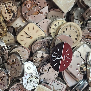 K&ouml;nnte beinhalten: Nahaufnahme einer Sammlung von Vintage-Uhrenteilen, darunter Zifferbl&auml;tter, Zahnr&auml;der und Mechanismen. Die Zifferbl&auml;tter haben verschiedene Formen, mit r&ouml;mischen und arabischen Ziffern. Die Zahnr&auml;der und Mechanismen sind aus Metall.