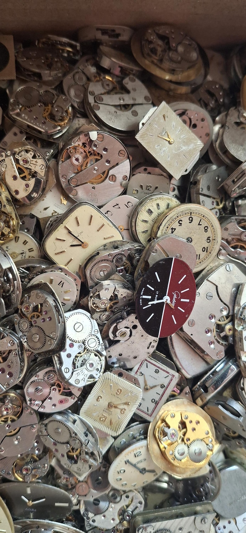K&ouml;nnte beinhalten: Nahaufnahme einer Sammlung von Vintage-Uhrenteilen, darunter Zifferbl&auml;tter, Zahnr&auml;der und Mechanismen. Die Zifferbl&auml;tter haben verschiedene Formen, mit r&ouml;mischen und arabischen Ziffern. Die Zahnr&auml;der und Mechanismen sind aus Metall.