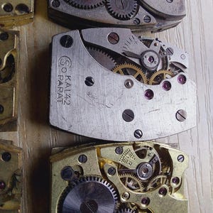Puede incluir: Primer plano de varios movimientos de reloj antiguos, que muestran intrincados engranajes, resortes y joyas. Los componentes met&aacute;licos son en tonos plateados y dorados, con peque&ntilde;as joyas de color rub&iacute;. El texto "SO KAL42 PARAT" es visible.
