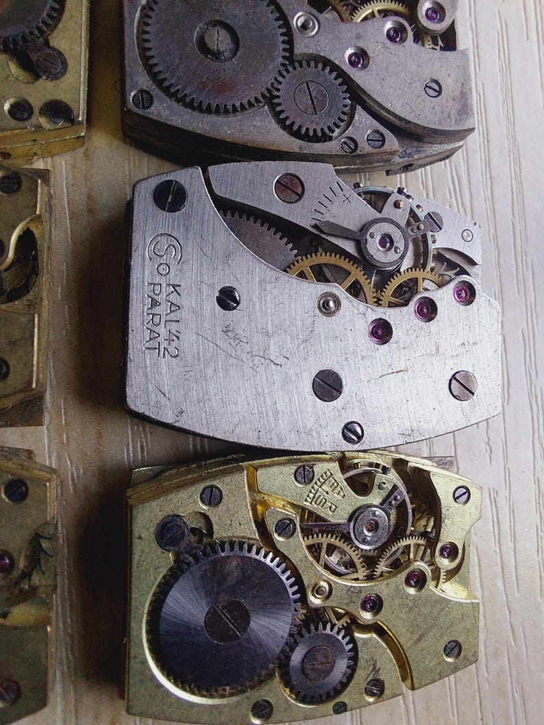 Puede incluir: Primer plano de varios movimientos de reloj antiguos, que muestran intrincados engranajes, resortes y joyas. Los componentes met&aacute;licos son en tonos plateados y dorados, con peque&ntilde;as joyas de color rub&iacute;. El texto "SO KAL42 PARAT" es visible.
