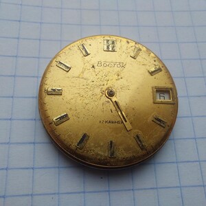 May include: A vintage gold-toned watch face with the word "Восток" in Cyrillic script. The face has rectangular hour markers, a date window at the 3 o'clock position, and two hands. The watch face is slightly worn.