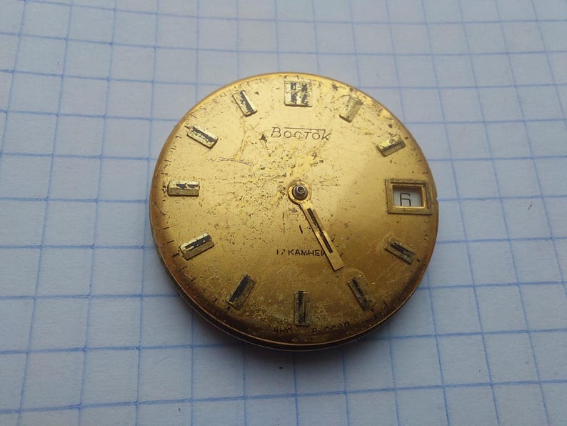 May include: A vintage gold-toned watch face with the word "Восток" in Cyrillic script. The face has rectangular hour markers, a date window at the 3 o'clock position, and two hands. The watch face is slightly worn.