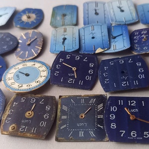Op de afbeelding: Een verzameling vintage wijzerplaten met blauwe en witte wijzerplaten. De wijzerplaten zijn in verschillende vormen en maten, en sommige hebben Romeinse cijfers. Het woord "Luch" is zichtbaar op verschillende van de wijzerplaten.