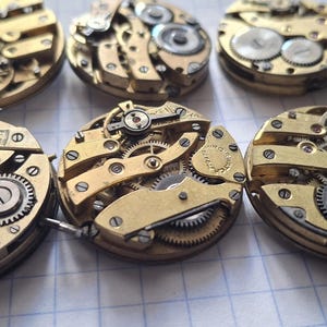 Op de afbeelding: Close-up van verschillende vintage uurwerkmechanismen, met ingewikkelde tandwielen, veren en andere mechanische componenten. De horlogeonderdelen zijn voornamelijk goudkleurig, met zilveren accenten, gerangschikt op een rasteroppervlak.