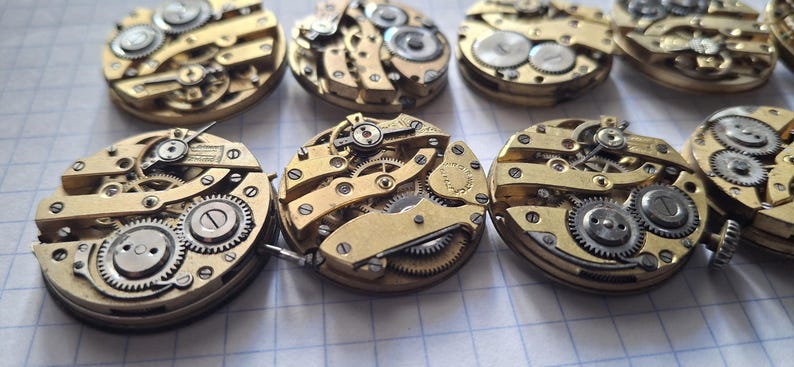 Op de afbeelding: Close-up van verschillende vintage uurwerkmechanismen, met ingewikkelde tandwielen, veren en andere mechanische componenten. De horlogeonderdelen zijn voornamelijk goudkleurig, met zilveren accenten, gerangschikt op een rasteroppervlak.