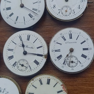 May include: Collection of antique watch faces with Roman numeral hour markers and sub-dials. The white faces show signs of age, with some featuring the names "Clairment" and "Le Boutte". The watch faces are arranged on a wooden surface.
