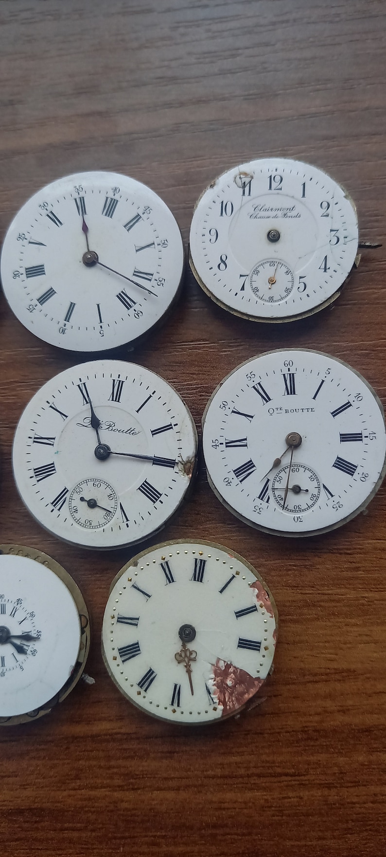 May include: Collection of antique watch faces with Roman numeral hour markers and sub-dials. The white faces show signs of age, with some featuring the names "Clairment" and "Le Boutte". The watch faces are arranged on a wooden surface.