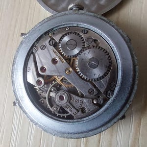 Puede incluir: Primer plano del mecanismo de un reloj vintage. La caja de metal plateado rodea los intrincados engranajes, muelles y joyas del funcionamiento interno del reloj. Las palabras "SWISS MADE" son visibles en una placa de metal. El reloj est&aacute; abierto.