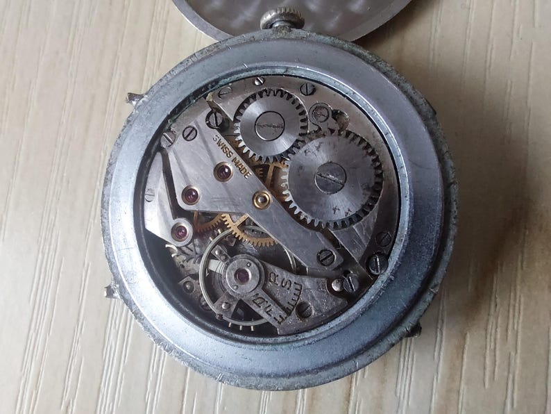Puede incluir: Primer plano del mecanismo de un reloj vintage. La caja de metal plateado rodea los intrincados engranajes, muelles y joyas del funcionamiento interno del reloj. Las palabras "SWISS MADE" son visibles en una placa de metal. El reloj est&aacute; abierto.