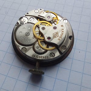 Puede incluir: Primer plano de un movimiento de reloj vintage con intrincados engranajes y componentes. El mecanismo plateado presenta engranajes dorados, un eje de cuerda y el n&uacute;mero "234305". El movimiento del reloj est&aacute; sobre un fondo de cuadr&iacute;cula azul.