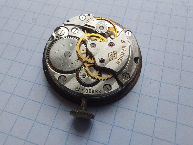 Puede incluir: Primer plano de un movimiento de reloj vintage con intrincados engranajes y componentes. El mecanismo plateado presenta engranajes dorados, un eje de cuerda y el n&uacute;mero "234305". El movimiento del reloj est&aacute; sobre un fondo de cuadr&iacute;cula azul.