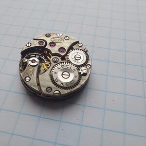 May include: Close-up of a vintage watch movement with intricate gears and components. The watch mechanism features the brand name "DOXA" and "LOCLE" etched on the metal. The gears are silver with pink jewels.