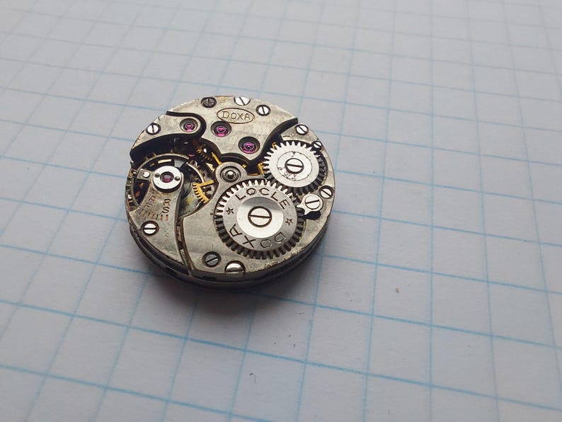 May include: Close-up of a vintage watch movement with intricate gears and components. The watch mechanism features the brand name "DOXA" and "LOCLE" etched on the metal. The gears are silver with pink jewels.