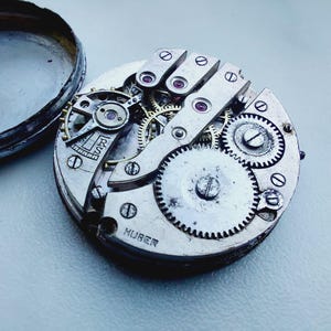 May include: A silver-toned mechanical pocket watch movement with an exposed gear train, balance wheel, and bridges. The metal plate is engraved with the text RS and HUBER. The circular mechanism is shown next to its detached, dark-colored metal case back.