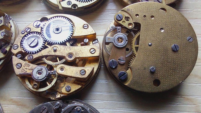 May include: Close-up of several antique watch movements, showcasing intricate gears, screws, and metal components. The watch parts are in various stages of disassembly, revealing the internal mechanisms. The brass and silver-colored elements are visible.