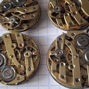 Op de afbeelding: Vier vintage uurwerkbewegingen met ingewikkelde tandwielen en mechanismen. De componenten zijn voornamelijk goudkleurig metaal met zilverkleurige tandwielen en schroeven. De horlogeonderdelen zijn gerangschikt op een witte geruite papieren achtergrond.