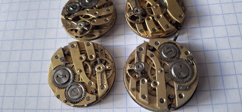 Op de afbeelding: Vier vintage uurwerkbewegingen met ingewikkelde tandwielen en mechanismen. De componenten zijn voornamelijk goudkleurig metaal met zilverkleurige tandwielen en schroeven. De horlogeonderdelen zijn gerangschikt op een witte geruite papieren achtergrond.