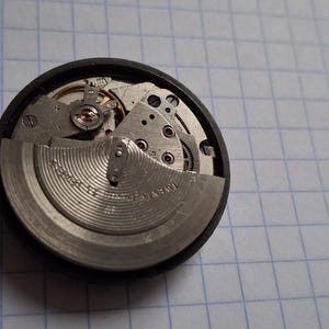 Puede incluir: Primer plano de un mecanismo de reloj, mostrando engranajes intrincados y un volante. Los componentes metálicos plateados y dorados están alojados en un marco circular negro. La imagen destaca la precisión de la ingeniería relojera.